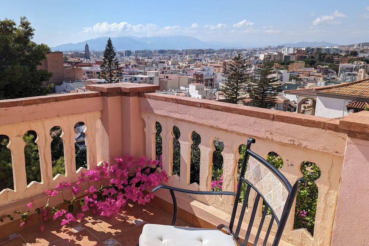 A balcony with a chair and pink flowers, overlooking a cityscape with buildings and mountains.