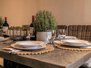 Une table à manger dressée avec des assiettes, des verres et une bouteille de vin, entourée d’un canapé en osier et d’une plante en pot.