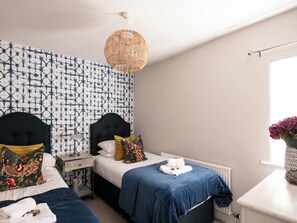 Un dormitorio con dos camas, una lámpara de techo de ratán, papel tapiz estampado y una ventana con un jarrón de flores.