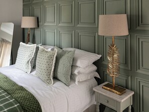 Een net opgemaakt bed met gestreepte kussens, een groen gebreid plaid en een nachtkastje met een gouden lamp.