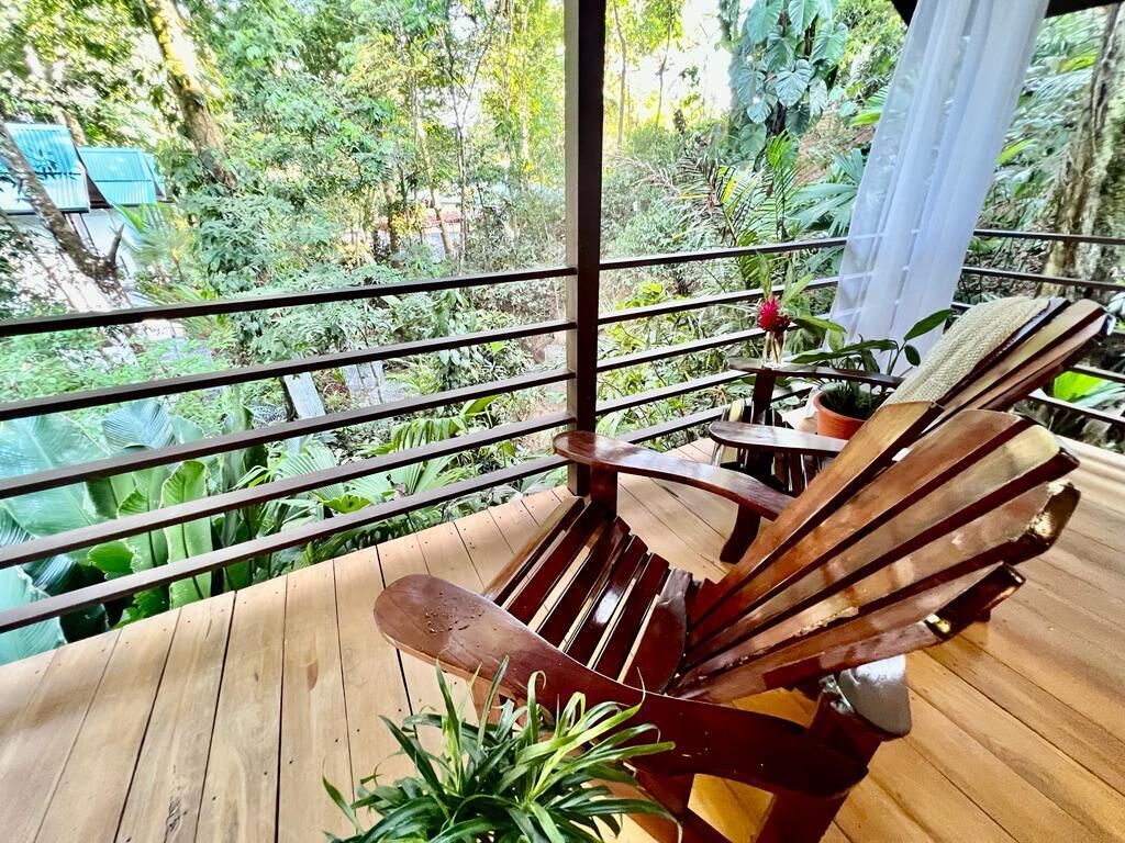 Une terrasse en bois, agrémentée d’une chaise et d’une table en bois, entourée de rampes en métal, et offrant une vue sur un paysage verdoyant et luxuriant.