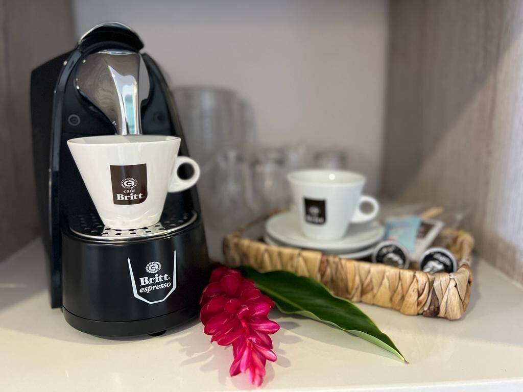Une machine à café avec une tasse et une soucoupe, un petit panier contenant des dosettes de café et une fleur rouge sur une table.