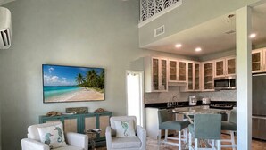 A living room with two armchairs, a coffee table, and a TV displaying a beach scene. Adjacent is a kitchen with a dining area. - Sand Dollar W212 Bayfront Escape- Gorgeous Sunset Views, Bikes Included! (Pensacola Beach)
