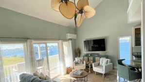 A spacious living room with a large sofa, two armchairs, a coffee table, and a dining area. A ceiling fan and a TV are mounted on the wall. The room has large windows with a view of the sea. - Sand Dollar W212 Bayfront Escape- Gorgeous Sunset Views, Bikes Included! (Pensacola Beach)