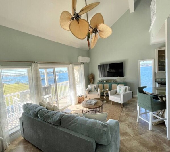 A spacious living room with a large sofa, two armchairs, a coffee table, and a dining area. A ceiling fan and a TV are mounted on the wall. The room has large windows with a view of the sea. - Sand Dollar W212 Bayfront Escape- Gorgeous Sunset Views, Bikes Included! (Pensacola Beach)