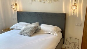 Una cama bien hecha con ropa blanca y un cabecero gris, un adorno en la pared encima y dos lámparas de pared a cada lado.
