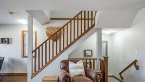 A spacious living room with a brown leather sofa, a wooden staircase, and a woven basket on the floor. - Downtown Basecamp | Stylish | Games Room (Invermere)