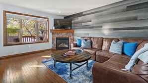 A modern living room with a stone fireplace, leather sofa, and a large window with a view of trees. - Downtown Basecamp | Stylish | Games Room (Invermere)
