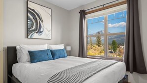 A modern hotel room with a large bed, a view of a mountain, and a painting on the wall. - Downtown Basecamp | Stylish | Games Room (Invermere)