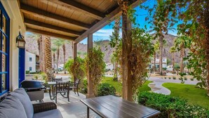 An outdoor patio with a sofa, table, and grill, surrounded by greenery and a view of a residential area. - Mountain Vista A | 1BR Legacy Villa (La Quinta)