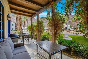 An outdoor patio with a sofa, table, and grill, surrounded by greenery and a view of a residential area. - Mountain Vista A | 1BR Legacy Villa (La Quinta)