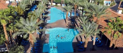 An aerial view of a resort with a central swimming pool surrounded by palm trees and lounge chairs.