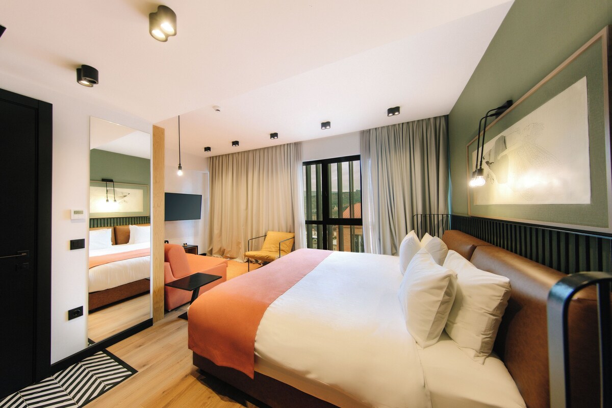A hotel room with a large bed, a seating area with a chair and table, a view of the outdoors through the window, and a painting on the wall.