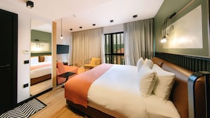 A hotel room with a large bed, a seating area with a chair and table, a view of the outdoors through the window, and a painting on the wall. - Hotel 21 (Tbilisi)