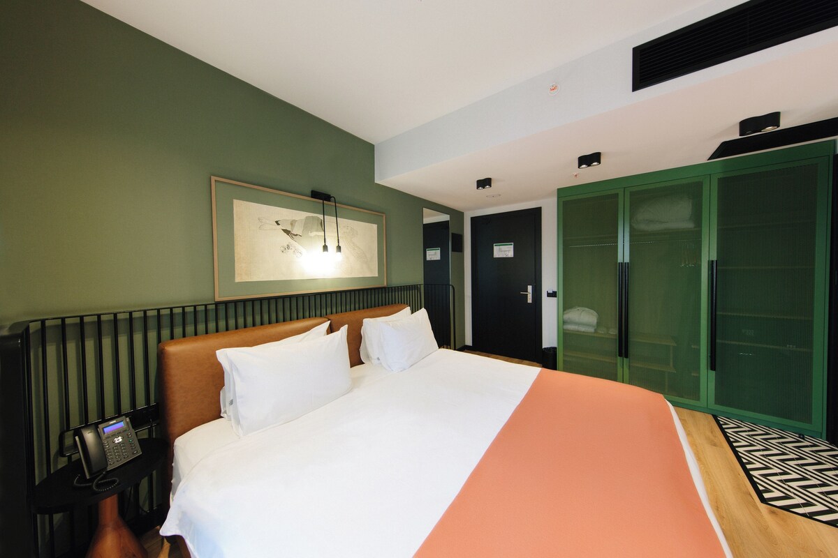 A hotel room with a bed, a telephone, a green wardrobe, and a framed picture on the wall.