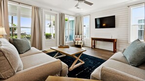 A spacious living room with a ceiling fan, large windows, a flat-screen TV, and a view of the outdoors. - New 3 Bedroom Beachfront Rental in downtown Bay St Louis (Bay St. Louis)