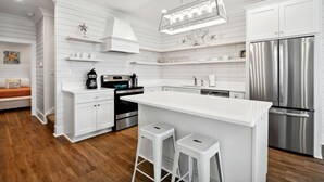 A modern kitchen with white cabinetry, a stainless steel refrigerator, a built-in oven, and a large island with stools. - New 3 Bedroom Beachfront Rental in downtown Bay St Louis (Bay St. Louis)