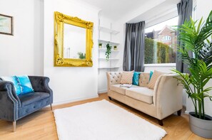 Una sala de estar con un sofá blanco, un sillón oscuro, un espejo grande con marco dorado y una planta en maceta.