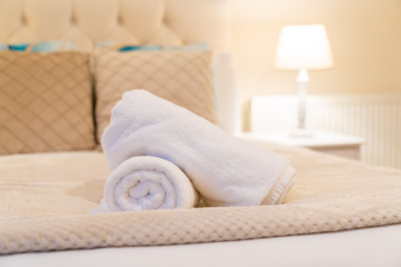 Una cama bien hecha con una toalla blanca enrollada, un cabecero acolchado color beige y una mesita de noche con una lámpara.