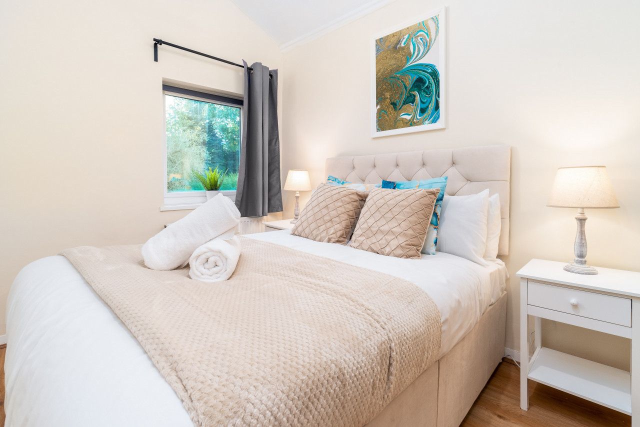 Un dormitorio con una cama grande, una ventana con cortinas, una mesita de noche con una lámpara y un cuadro en la pared.