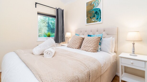 Un dormitorio con una cama grande, una ventana con cortinas, una mesita de noche con una lámpara y un cuadro en la pared.