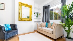 A living room with a white sofa, a dark armchair, a large mirror with a gold frame, and a potted plant.