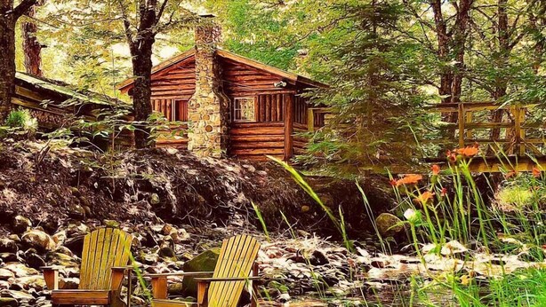 Una cabaña rústica con chimenea de piedra, rodeada de un bosque denso y un pequeño arroyo. Dos sillas Adirondack están situadas cerca de la orilla.