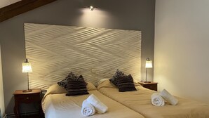 Habitación de hotel con un gran cabecero texturizado, dos camas con ropa de cama color beige, dos lámparas de noche y una cama bien hecha con una toalla enrollada.