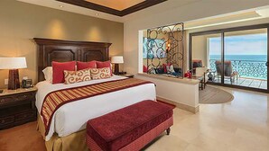 A spacious bedroom with a large bed, a red bench, a nightstand, and a view of the ocean through a balcony.