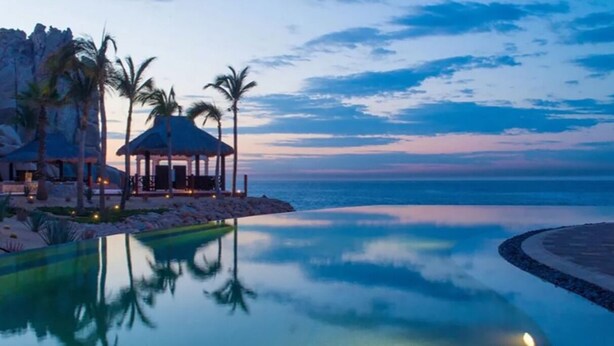 A resort with a clear pool, palm trees, and a gazebo overlooking the ocean at dusk.