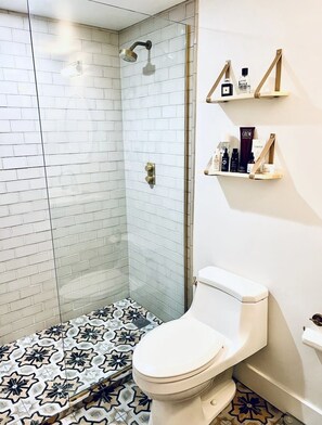 Une salle de bain moderne avec une douche à l’italienne en verre, des toilettes posées sur un carrelage à motifs et une étagère contenant des articles de toilette.