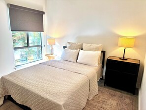 A bedroom with a bed, two bedside lamps, a window with a view of greenery, and a small wooden nightstand.