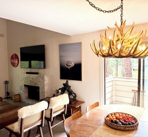 Un salon moderne doté d’une cheminée, d’un téléviseur fixé au mur, d’un lustre décoratif en forme de bois de cerf et d’un coin repas agrémenté d’un panier de fruits.
