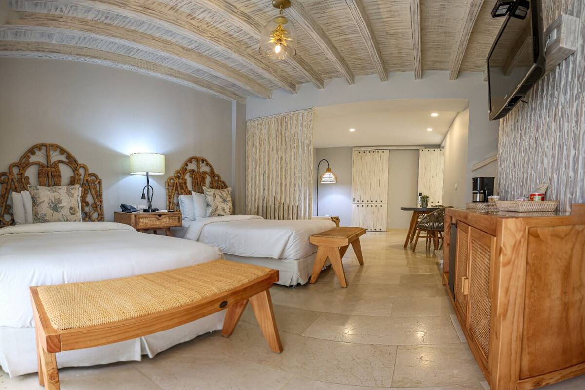 Habitación de hotel con dos camas, un banco de madera, un escritorio y un televisor montado en la pared.