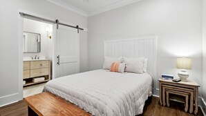 A bedroom with a bed, a bedside table with a lamp, a bench, a dresser, and a sliding door. - New 3 Bedroom Beachfront Rental in downtown Bay St. Louis (Bay St. Louis)