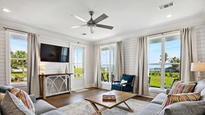 A spacious living room with a large flat-screen TV, a ceiling fan, and a view of a lush garden through large windows. - New 3 Bedroom Beachfront Rental in downtown Bay St. Louis (Bay St. Louis)