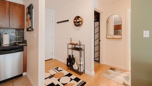 A modern kitchen with wooden cabinets, a dishwasher, and a tiled backsplash. There is a hallway with a black and white patterned rug, a small shelf with decorative items, and a mirror on the wall.