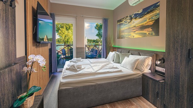 A bedroom with a bed, a TV, a painting, and a view of the outdoors. - Mártoni Resort & Restaurant (Szigetszentmarton)