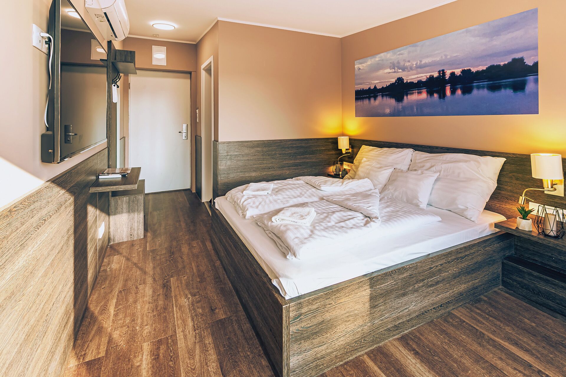 A hotel room with a wooden bed, a nightstand with a lamp, a painting of a lake, and a wooden floor.