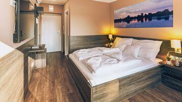 A hotel room with a wooden bed, a nightstand with a lamp, a painting of a lake, and a wooden floor.