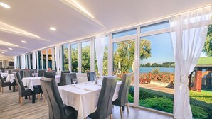 A restaurant with tables set for dining, large windows offering a view of a lake and greenery. - Mártoni Resort & Restaurant (Szigetszentmarton)