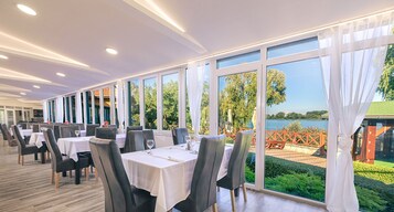 A restaurant with tables set for dining, large windows offering a view of a lake and greenery.