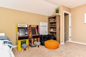 A bedroom with two arcade machines, a bean bag, a small stool, a door, and a shelf with a plant.