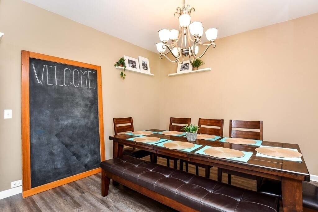 A dining room with a wooden table, four chairs, a bench, a chandelier, and a 'WELCOME' sign on the wall.