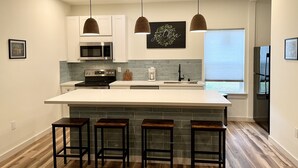 A modern kitchen with a central island, bar stools, a microwave, a coffee maker, and pendant lighting. - Hidden Bungalow 1 1/2 blocks from the beach (Bay St. Louis)