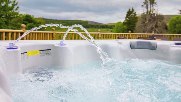 A hot tub with water jets, surrounded by a wooden deck and a scenic view of hills and trees. - Dalnaglar Cottage - UK30260 (Glenshee, Cairngorms)