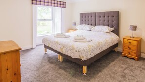 A bedroom with a large bed, two bedside tables, a wooden dresser, a window with a checkered valance, and a ceiling light. - Dalnaglar Cottage - UK30260 (Glenshee, Cairngorms)