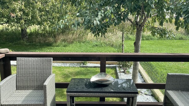 Una terraza de madera con mobiliario de mimbre, una mesa de vidrio y vistas a un área verde con árboles.