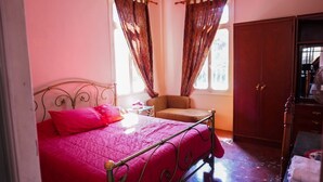 Un dormitorio con estructura de cama metálica, cubrecama rosa, una silla y un armario.