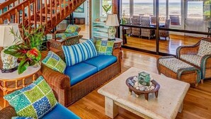 A spacious living room with a wooden staircase, wicker furniture, and a view of the beach. - B.I. Kaiakea,  Sleeps 12, Hot Tub, Astounding Oceanfront in Tranquil Puako! (Kamuela)